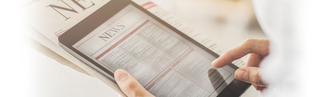 A person's hand is holding a tablet displaying a newspaper with the word "NEWS" prominently featured. The tablet screen shows sections of text, suggesting digital news reading. In the background, a physical newspaper is partially visible.