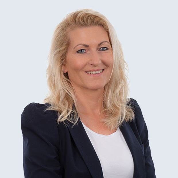 A woman with long, light blonde hair smiles while wearing a black blazer over a white top. She poses against a light blue background, conveying a professional and approachable demeanor.