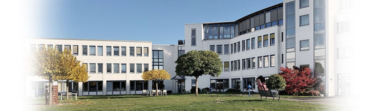 A modern building complex featuring multiple stories and large windows. In the foreground, there is a grassy area with trees and benches. The scene is set under a clear blue sky, indicating pleasant weather.
