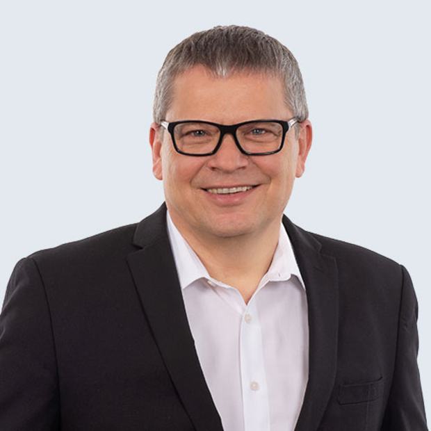 A middle-aged man with short gray hair and glasses smiles confidently. He wears a black suit jacket over a white shirt. The background is light blue, emphasizing his friendly demeanor.