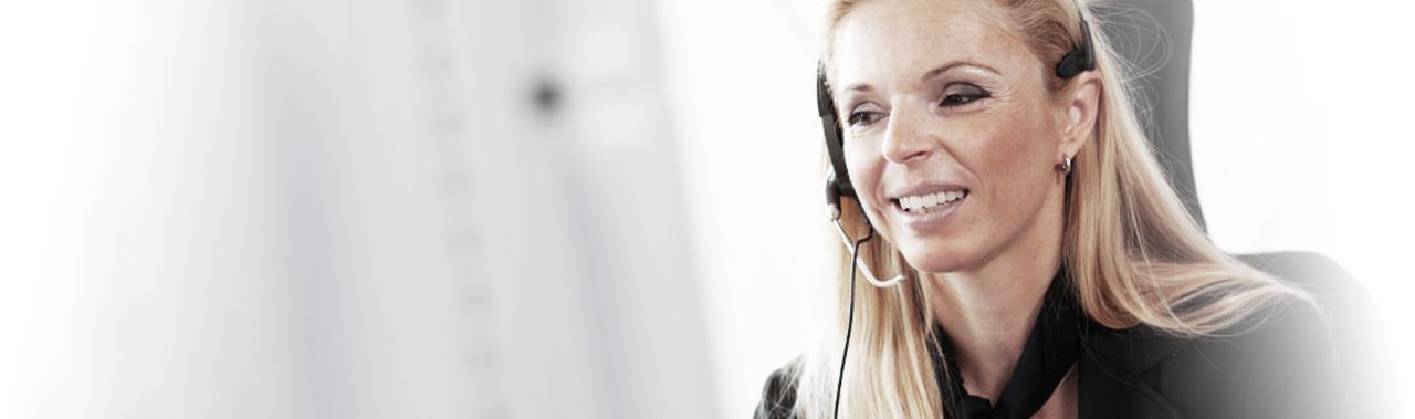 A smiling woman is wearing a headset and engaged in conversation, appearing focused and professional. She is sitting in front of a computer screen, suggesting a work environment related to customer service or communication. The background is softly blurred to emphasize her expression.